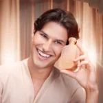 Freedom Musk Bouquet | 27 smiling man holding the bottle beside his face, warm curtain backdrop