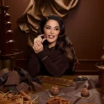 Woman holding a piece of chocolate near her face at a table with chocolate pieces and a chocolate fountain