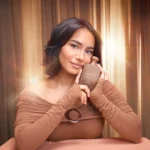 Freedom Musk Latte | 41 woman in a brown off-shoulder top holding the bottle against her cheek, warm curtain backdrop